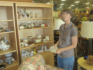 Woman displaying a decorative plate beside a shelf of pottery and figurines